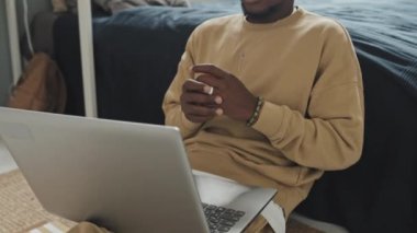 Kablosuz kulaklıklı Afro-Amerikan bir adamın yatak odasında otururken ve bilgisayardaki video görüşmesinde sohbet ederken görüntüsünü kaldır.