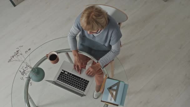 从上往下拍摄的老年妇女在笔记本电脑上打字 而坐在玻璃桌旁 带着抄本 咖啡和鲜花花瓶 从家庭和远程教育概念出发开展工作 — 图库视频影像