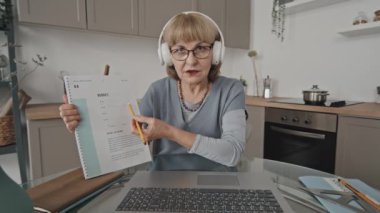 Kablosuz kulaklık takan kıdemli bayan öğretmen evden online ders verirken çalışma kitabındaki görevi açıklıyordu.