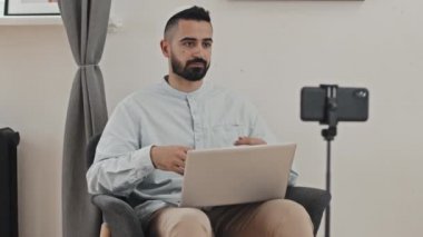 Kufi 'deki Müslüman bir adam evinde dizüstü bilgisayarla oturmuş akıllı telefon kamerasına bakıyor ve evden ders verirken ona dersini anlatıyor.