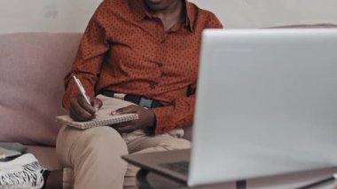 Afro-Amerikalı bir kadının evdeki koltukta oturup dizüstü bilgisayardan ders izlerken not defterine bir şeyler yazdığı orta bölüm fotoğrafı.