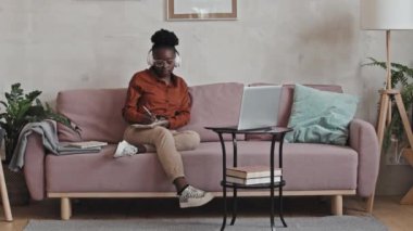 Kablosuz kulaklıklı Afro-Amerikalı bir kadın evdeki koltukta oturuyor, bilgisayardan online ders izliyor ve uzaktan çalışırken notlar alıyor.