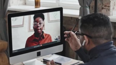 Ortadoğulu kablosuz kulaklıklı bir adam masada oturuyor ve bilgisayar üzerinden Afrikalı Amerikalı bir kadınla sohbet ediyor.