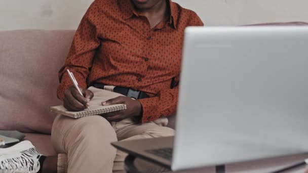 美国非洲裔妇女坐在家中沙发上 一边在笔记本上看在线课程 一边在笔记本上写下一些东西的中间截图 — 图库视频影像