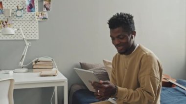 Kablosuz kulaklıklı Afro-Amerikalı bir adam yatak odasında oturmuş dijital tabletten online video görüşmeleri yapıyor.