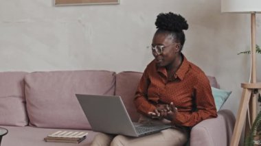 Neşeli Afrikalı Amerikalı kadın evdeki kanepede oturuyor, internette çalışırken el sallıyor, gülümsüyor ve sohbet ediyor.