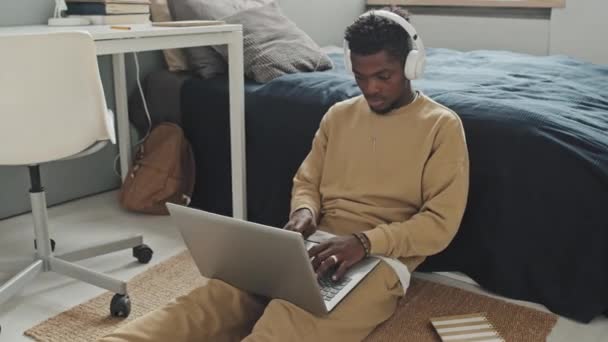 非洲裔美国人 戴着无线耳机 坐在卧室地板上 在笔记本电脑上打字 同时在离家很远的地方工作 — 图库视频影像