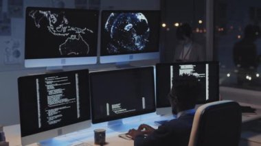 Genç siyahi erkek programcının omzunun üstünde geceleri şirket ofisinde beş monitörün önünde oturan bilgisayar kodu üzerinde çalışan, daktilo yazan, arka planda duran Asyalı kadın.