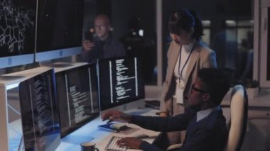 Orta uzunlukta, resmi kıyafetler giyen genç Asyalı bir kadın, siyah erkek bilişim uzmanıyla konuşuyor, o da geceleri masada oturuyor, şirket ofisinde bilgisayar kullanıyor.