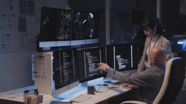 Orta uzunluktaki Asyalı genç bayan yönetici, beyaz erkek programcıyla konuşuyor, o da geceleri şirket ofisinde bilgisayar kullanıyor, siyahi adam telefonda arka planda konuşuyor.