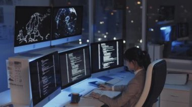 Genç Asyalı kadın programcının yan görüntüsü. Geceleri şirket ofisinde oturuyor, kodluyor, birden fazla monitör kullanıyor.