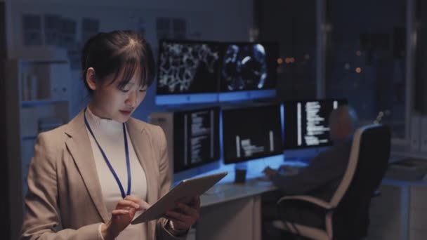 タブレットコンピュータを使用して 夜にオフィスに立っている若いアジアの実業家のウエストアップ 背景にぼやけたプログラマーのコーディング — ストック動画