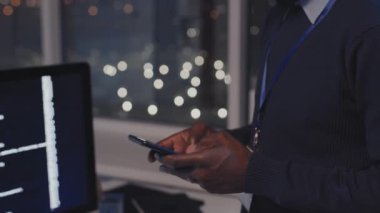 Kesilmiş erkek bilişim uzmanının yan görüntüsü modern ofiste gece ayakta duruyor, akıllı telefon kullanıyor, mesaj yazıyor.