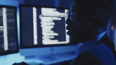 Genç siyahi erkek programcının karanlık kurumsal ofiste masada oturması klavyeye yazması, bilgisayarda kodlaması, birden fazla monitör kullanması.