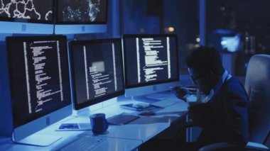 Genç siyahi erkek programcının karanlık kurumsal ofiste masa başında oturup bilgisayarların çalışmasını izlemesi, gece karton kutudan şehriye yemesi.