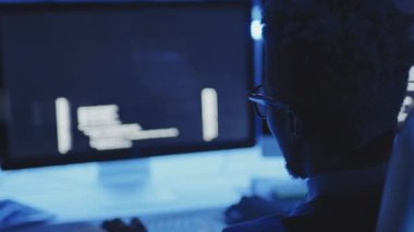 Karanlık kurumsal ofiste masa başında oturan, klavyede yazan, bilgisayarda kodlayan genç siyahi erkek programcının omzunun üstünden.