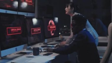 Kritik hatayı düzelten iki genç siber güvenlik uzmanının yan görüntüsü. Geceleri masadaki bilgisayar monitörlerinde uyarı alarmı.
