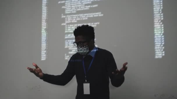 ガラスとカスタムメイドのフェイスマスクを身に着けている精力的な若い黒の男性ソフトウェアエンジニアの中 それにコンピュータコードの投影と白い壁に立って プレゼンテーションを作る — ストック動画
