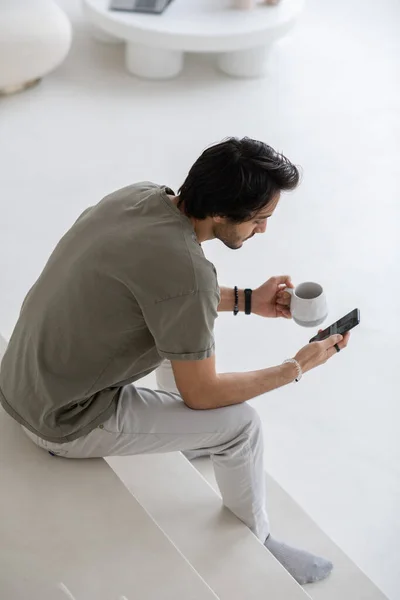 Młody nowoczesny menedżer z telefonem komórkowym siedzącym na białych schodach i SMS-em — Zdjęcie stockowe