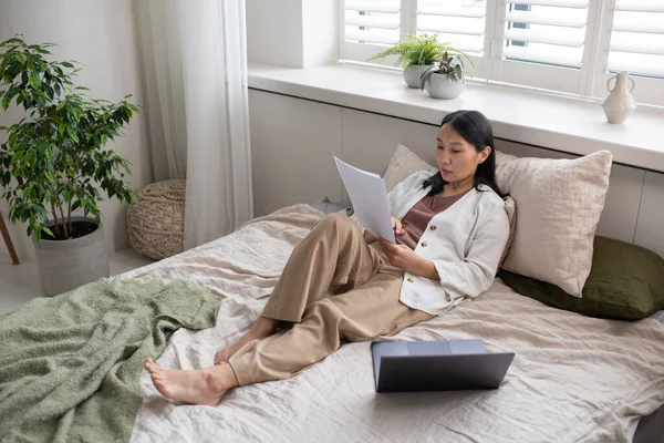 Молодая уверенная азиатка в повседневной одежде читает газеты на кровати — стоковое фото