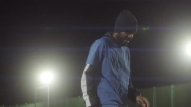Üniforma giyen genç Afrikalı Amerikalı erkek sporcunun eğilip topla futbol hareketleri çalışması, geceleri spot ışıkları altında futbol sahasında durması.