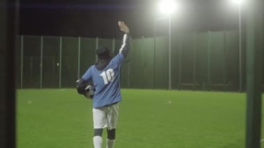 Genç siyah bir adam spor kıyafetleri ve kalın bir şapka takıyor, topu tutuyor, boş futbol sahasına giriyor, görünmez seyirciyi selamlıyor, ünlü bir oyuncu olmanın hayalini kuruyor.