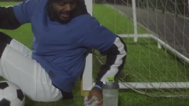 兴奋得上气不接下气的黑人 身穿制服 头戴暖和的帽子 坐在球场上的足球球门旁边 晚上在足球场上训练后喝水 — 图库视频影像
