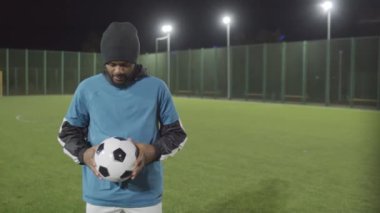 Spor kıyafetleri giyen, futbol topu tutan, futbol sahasının ortasında duran, başını kaldırıp kameraya bakan amatör siyah erkek sporcu.