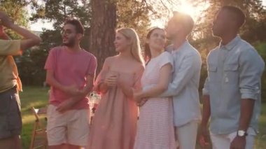 Group of modern young men and women spending time together standing against dining table in park having fun chatting, and dancing