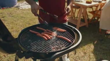 Barbeküde duran tanınmamış adam yaz günü açık hava partisi için sosis pişiriyor.