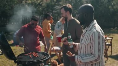 Genç Siyahi ve Kafkasyalı erkekler dışarıda bbq partisinde eğlenirken arkadaşları sosis pişirirken ve masa servisi yaparken bira içiyorlar.