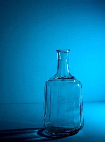 Glass empty open bottle with copy space. Graphic still life with light and shadow in blue light. The concept of glass containers, recyclable materials or alcoholism. Large creative vase. Open flask.