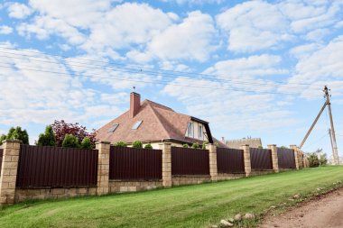Brown metal profile fence with block posts. Incline construction. Corrugated surface. Security. Private property fencing. Opaque hedge. Outdoor house exterior. Side view. Urban or industrial style.