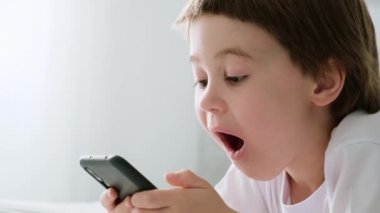 4 bin. 5 yaşındaki küçük oğlan akıllı telefon oyunuyla yatıyor ve oynuyor. Vay canına. Yakın plan çocuk portresi. Cep telefonunda duruyor. Video görüntüleri. Online eğitim kavramı. Beyaz oda ve kıyafetler.