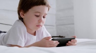 5 yaşındaki küçük çocuk yatağa uzanıyor ve akıllı telefonuyla oynuyor. Yakından bir portre. 4K video görüntüleri. Çevrimiçi eğitim kavramı. Beyaz oda ve kıyafetler. Çocuk telefon ekranını tutuyor ve kaydırıyor