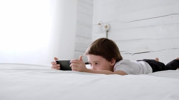Liten 5-årig pojke lägger sig på sängen och leker med smartphone. Närbild porträtt. 4K-videofilmer. Nätutbildningskoncept. Vita rummet och kläder. Barnet håller och bläddrar i telefonskärmen — Stockvideo