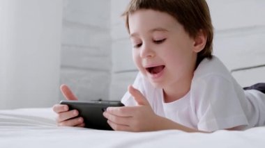 Mutlu 5 senelik çocuklar akıllı telefon video oyunu oynuyor. Çocuk oyun oynamak ya da öğrenmek için cep telefonu kullanıyor. Beyaz gömlekli gülümseyen çocuk yatak odasında yatıyor. Online eğitim. Yakın plan. 4K video görüntüleri. Boşluğu kopyala