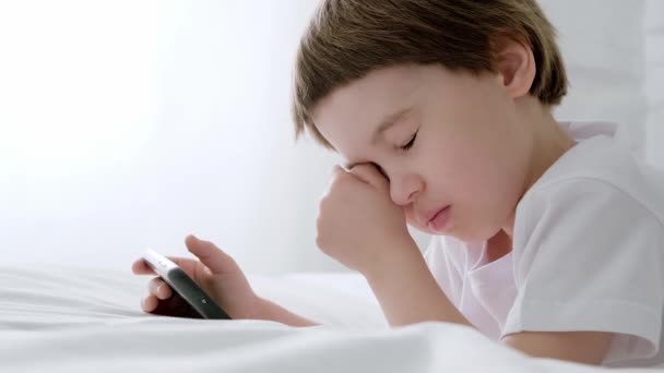 Mały 5-letni chłopiec leży na łóżku i bawi się grą na smartfonie. Dziecko jest zmęczone i chce spać. Masuje napięte oczy. Zbliżenie. Odbiór. Okulistyka. Zachowaj wzrok dziecka. Białe ubrania. Zbyt długo. — Wideo stockowe
