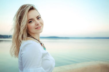 Genç Avrupalı kadın portresi ayakta, gülümseyen ve kameraya bakan. Çıplak makyaj malzemesi. Güzel bir yüz, büyük mavi gözler ve uzun sarı kıvırcık saçlar. Sabah tatili. Doğal deniz arka planı.