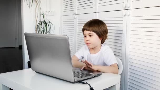 Niño de 5 años sentarse en la mesa, utiliza el ordenador portátil, mirando a la pantalla. El niño está haciendo la lección de deberes, jugando videojuegos, estudiando desarrollo de TI, viendo dibujos animados. Casa distancia concepto de educación en línea — Vídeos de Stock