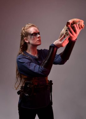 Siyah barbar fotografı olan, ortaçağ Viking kostümlü genç kızıl saçlı kadının yakın portresi. Kırmızı ışıklandırmalı stüdyo arka planına poz vermek için koyun kafatası tutmak.   