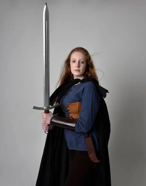Close up portrait of red haired woman wearing medieval viking inspired costume and flowing cloak,  Holding a long sword weapon, action pose against studio background.