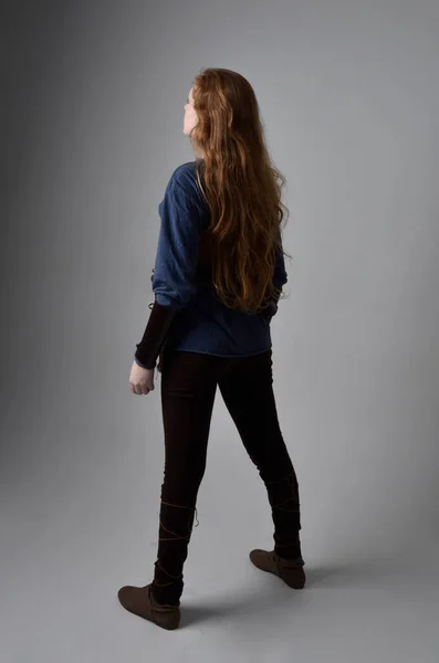 Full length portrait of red haired woman wearing medieval viking inspired costume,  standing pose with back to the camera, against studio background.