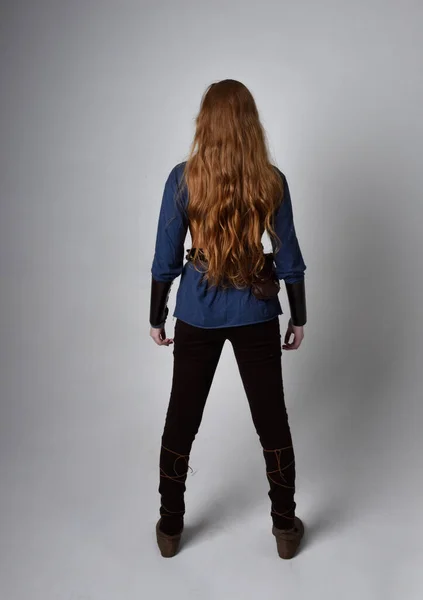 Full length portrait of red haired woman wearing medieval viking inspired costume,  standing pose with back to the camera, against studio background.