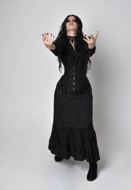 Siyah Viktorya dönemi korseli cadı kostümü giyen, stüdyo arka planında el hareketleriyle duran koyu renk saçlı kadının tam boy portresi..