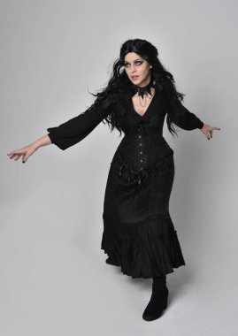 Siyah Viktorya dönemi korseli cadı kostümü giyen, stüdyo arka planında el hareketleriyle duran koyu renk saçlı kadının tam boy portresi..