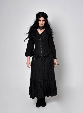 Siyah Viktorya dönemi korseli cadı kostümü giyen, stüdyo arka planında el hareketleriyle duran koyu renk saçlı kadının tam boy portresi..