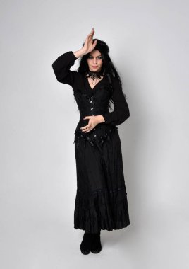 Siyah Viktorya dönemi korseli cadı kostümü giyen, stüdyo arka planında el hareketleriyle duran koyu renk saçlı kadının tam boy portresi..