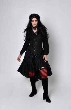 Siyah Viktorya dönemi korseli cadı kostümü giyen, stüdyo arka planında el hareketleriyle duran koyu renk saçlı kadının tam boy portresi..