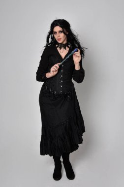 Siyah Viktorya dönemi korseli cadı kostümü giyen, stüdyo arka planında el hareketleriyle duran koyu renk saçlı kadının tam boy portresi..